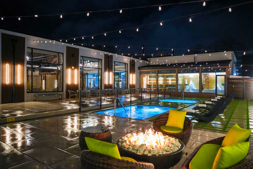 Outdoor pool courtyard at Fusion Spa & Resort in the evening, featuring illuminated pools, modern lounge seating, a central fire pit, and string lights creating a warm, inviting ambiance.